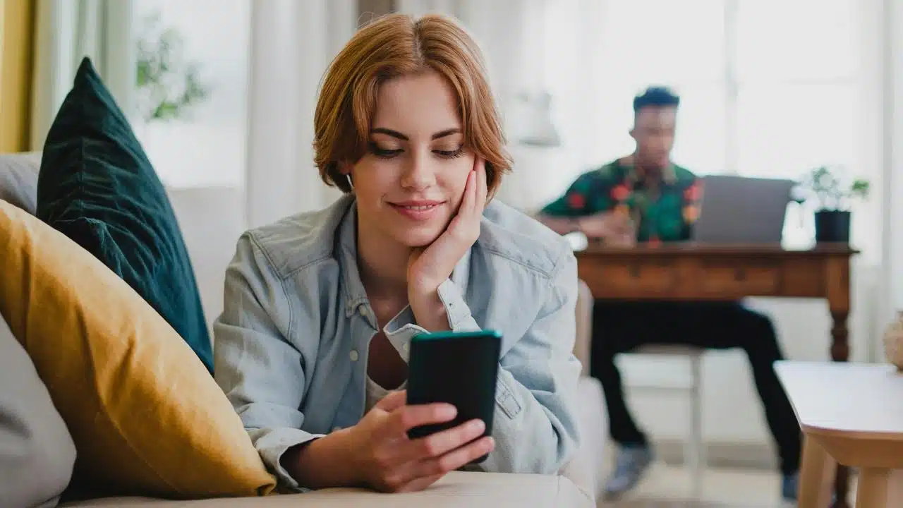A woman scrolling her phone on a couch while a man works at a desk behind her.