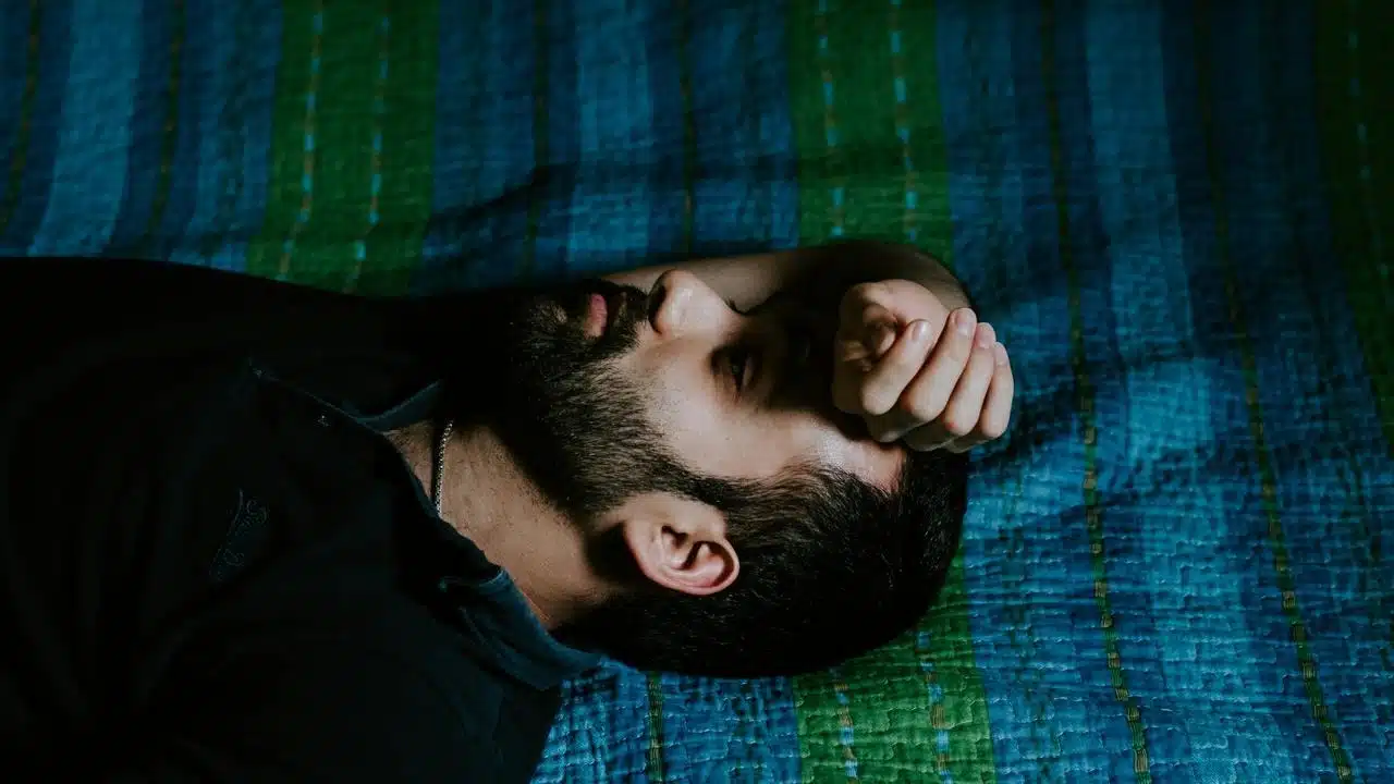 A bearded man lying on a bed with his hand resting on his forehead.
