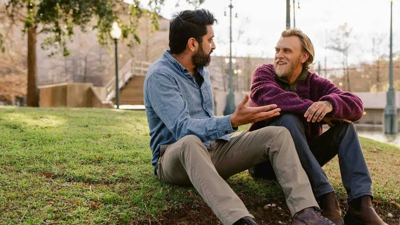 A man gesturing as he talks to another man while they sit on grass in a park.