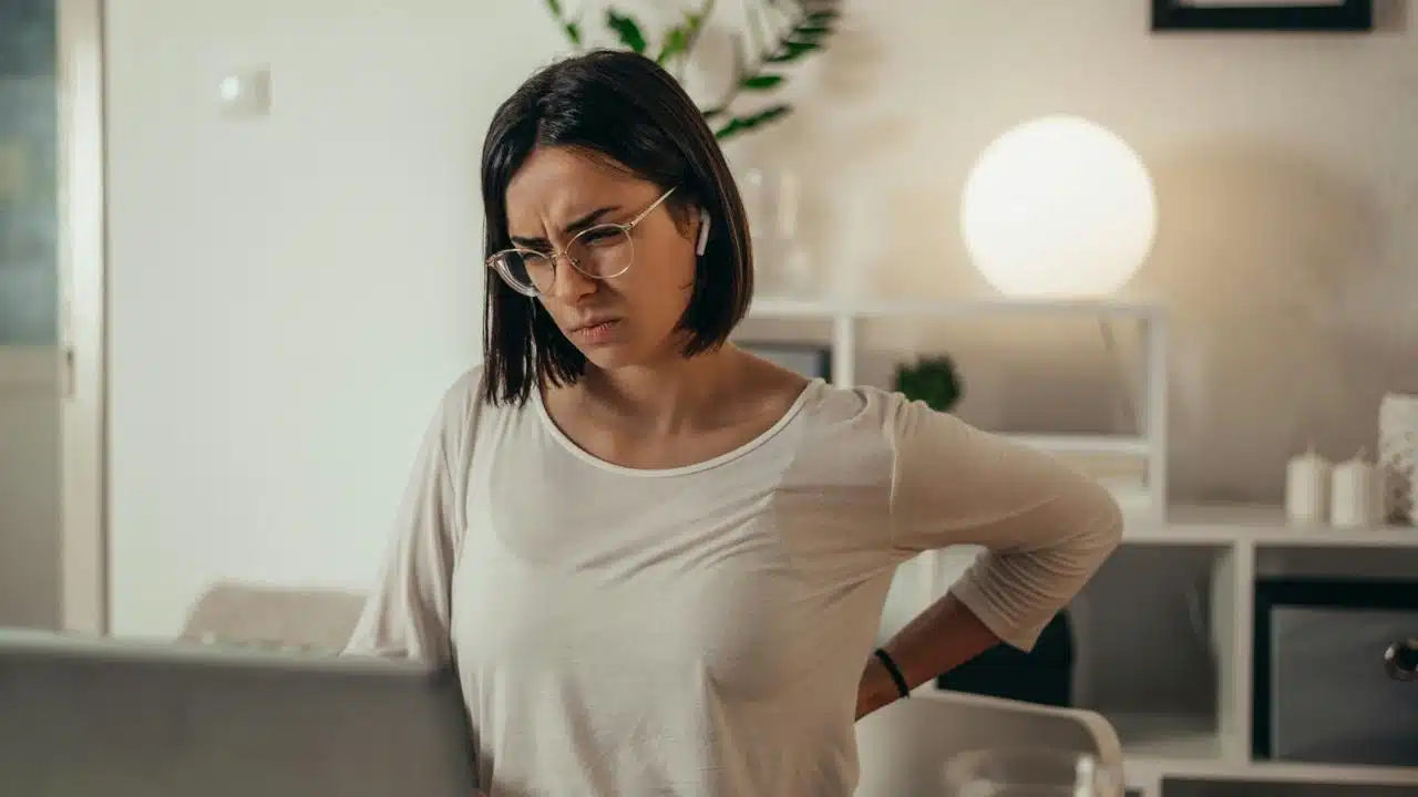 A woman clutching her lower back in discomfort while working on a laptop.