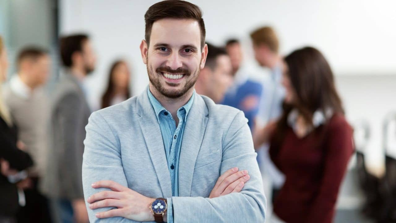 A confident man with his arms crossed and a friendly smile stands in a crowd of people.