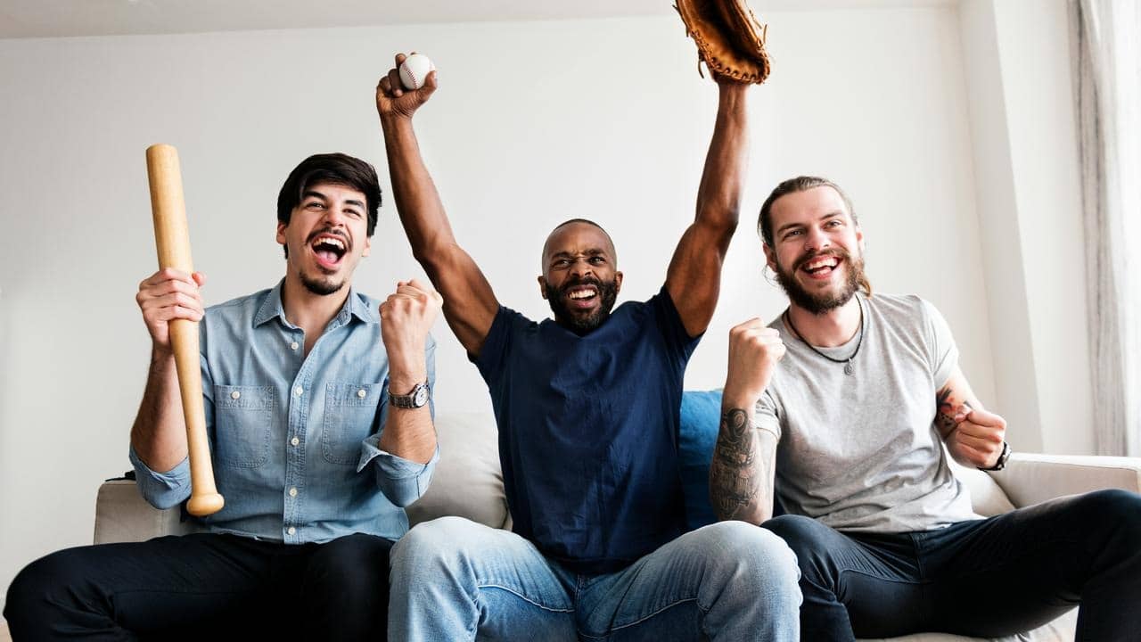 A trio of friends cheering on a couch with baseball gear.