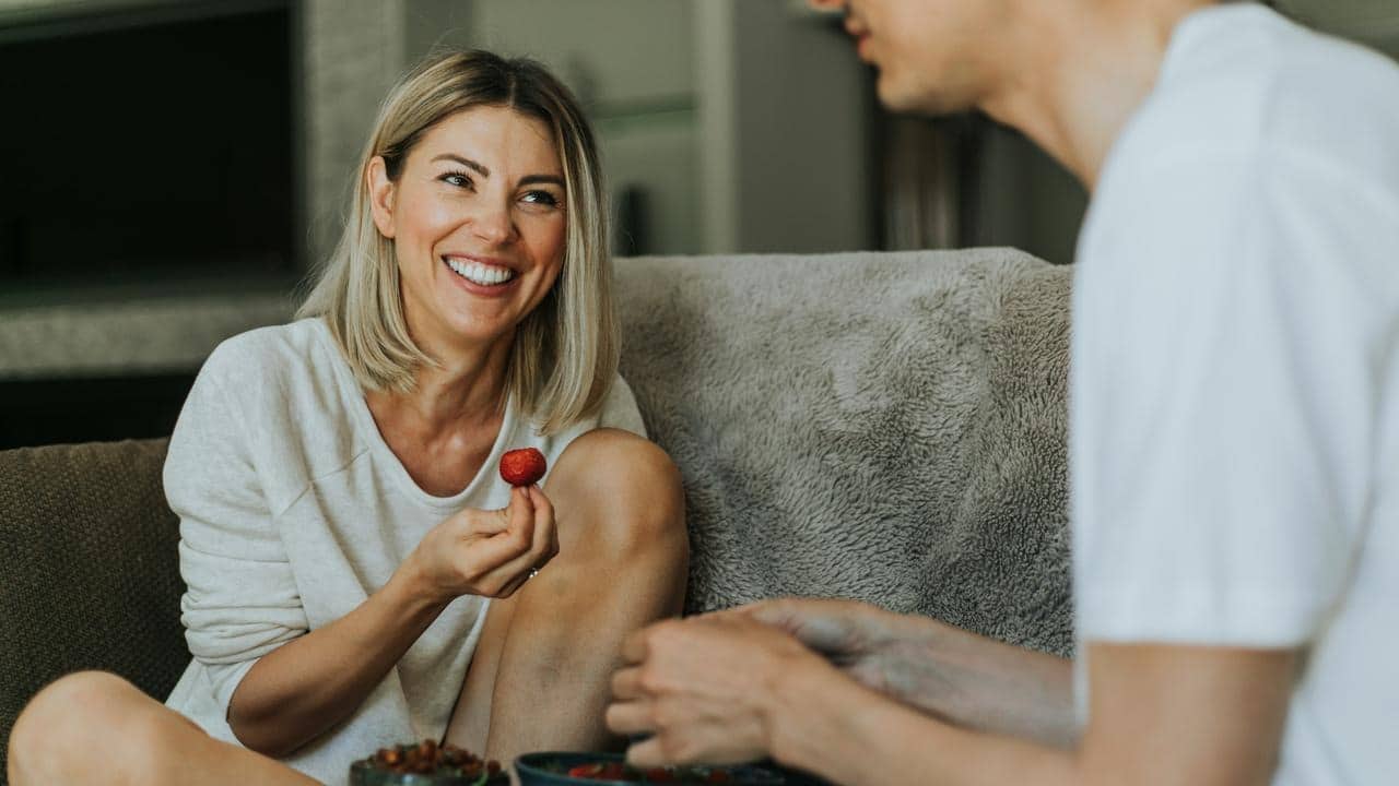 A woman smiling on a couch while holding a strawberry.