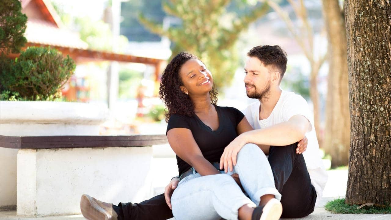 A couple sitting on the ground under a tree, smiling at each other.