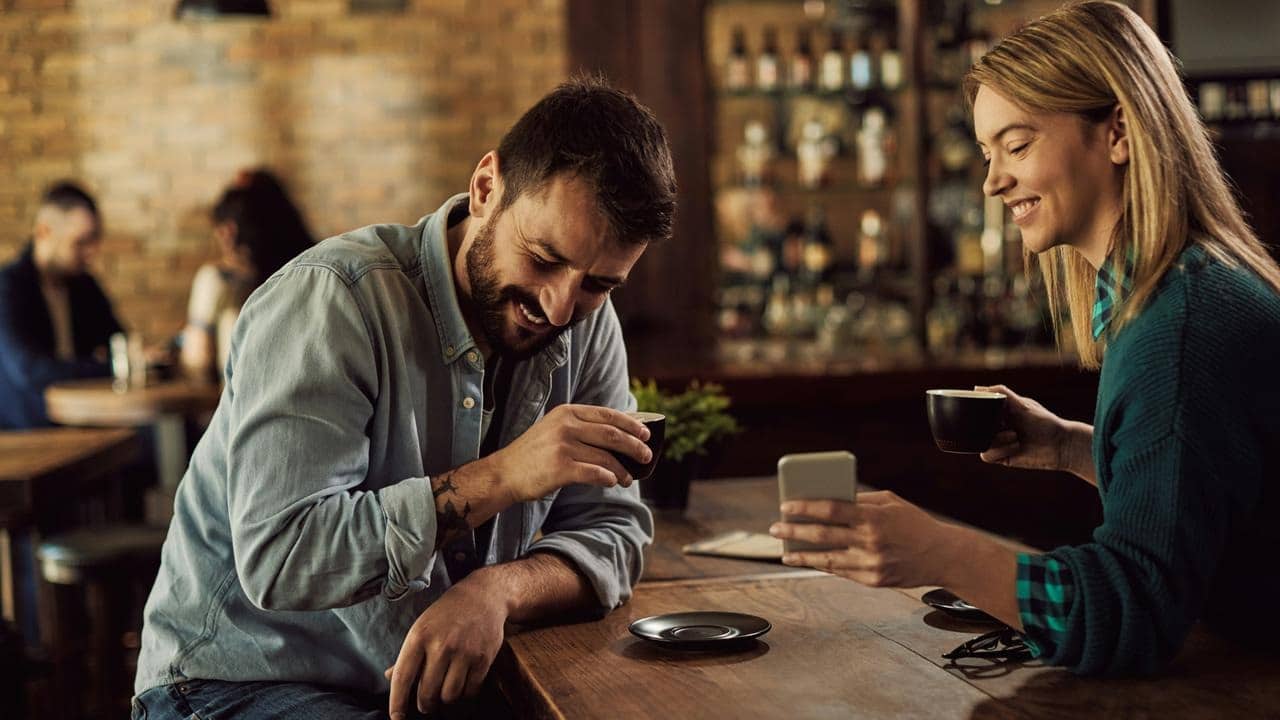 A man and woman sharing coffee while looking at a phone in a cafe.