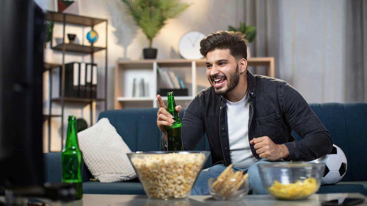 A man cheering on a couch with beer and snacks.