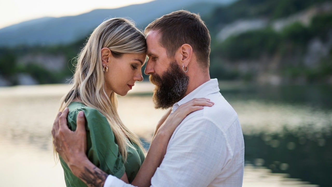 A bearded man touching foreheads with a blond woman outdoors.