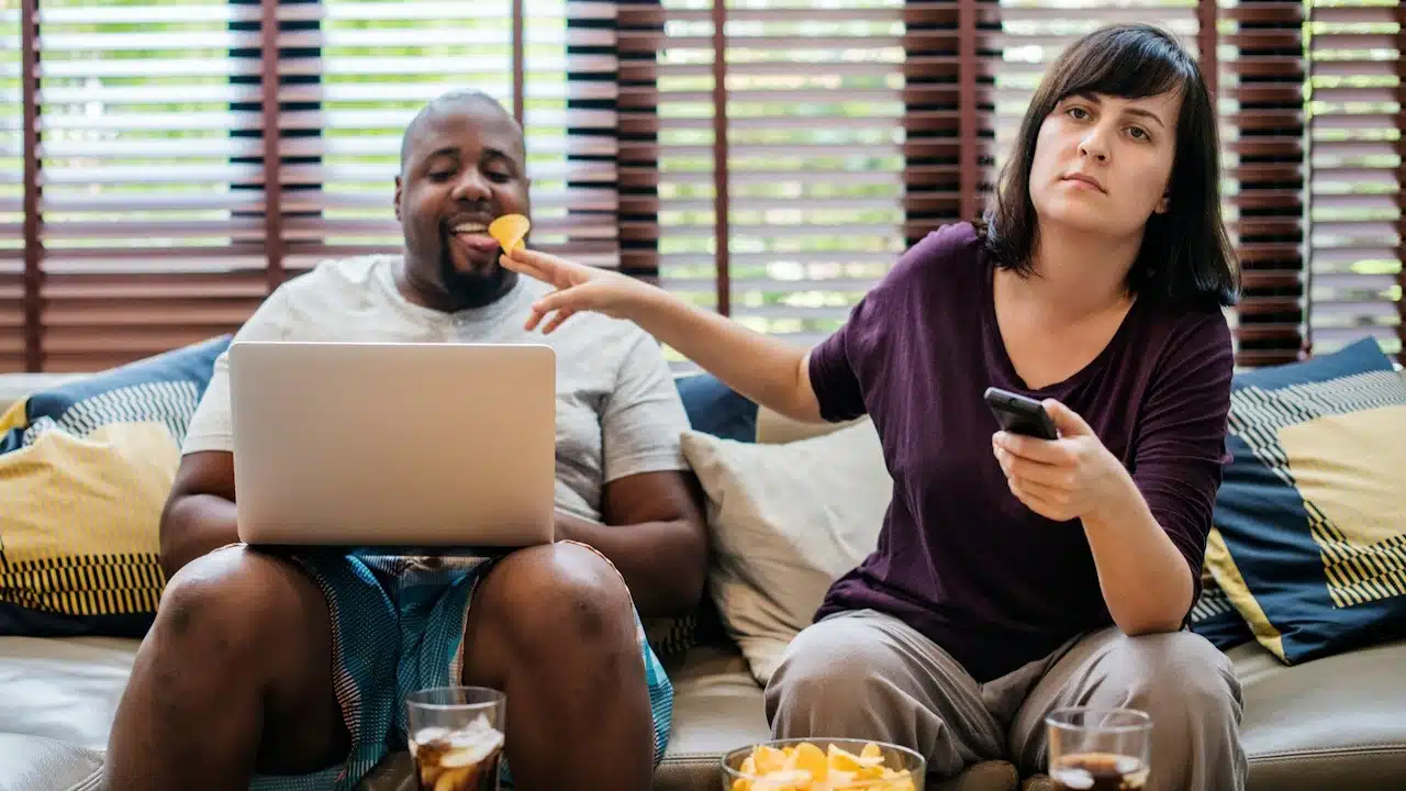 A man feeding a man chips while using a mobile phone and looking inscrutable.
