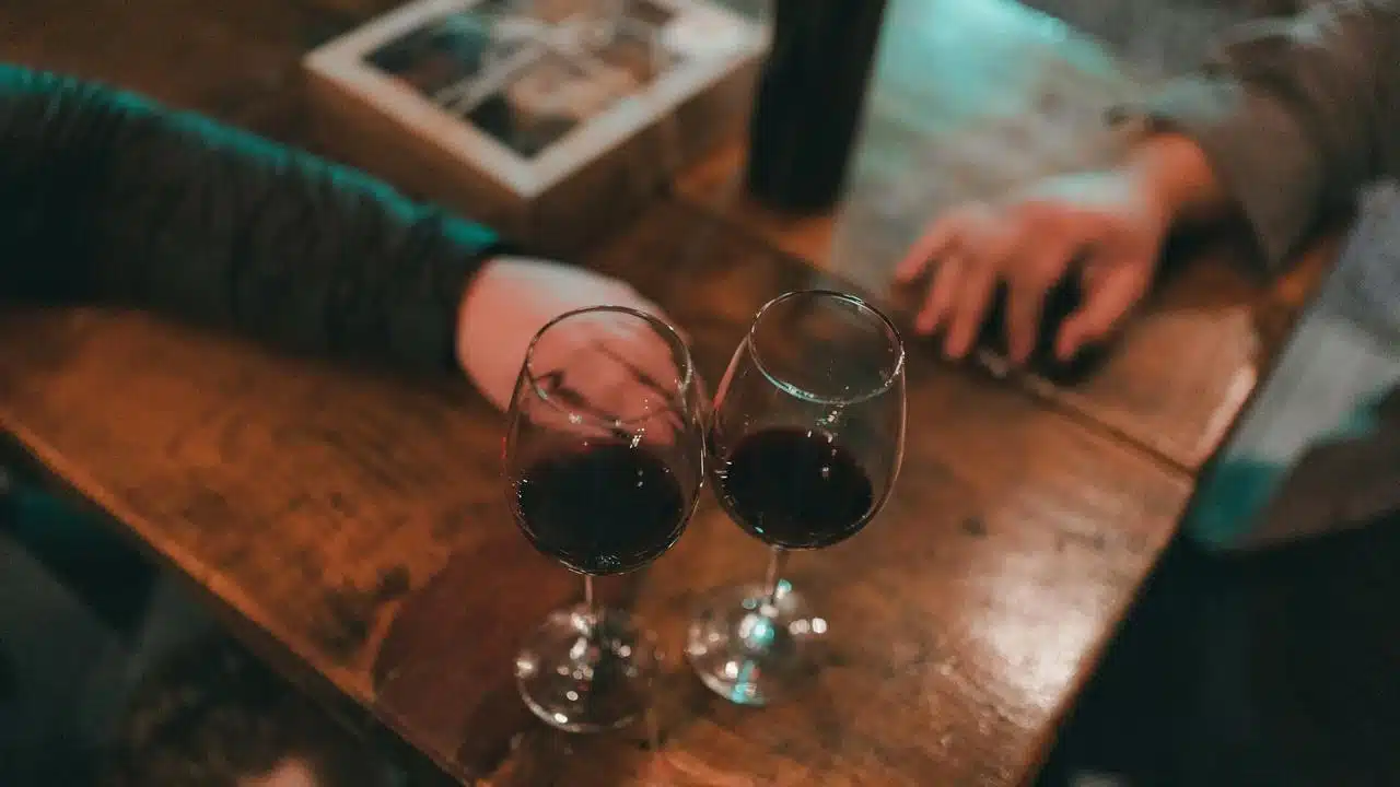 A pair of red wine glasses on a wooden table with two hands nearby.