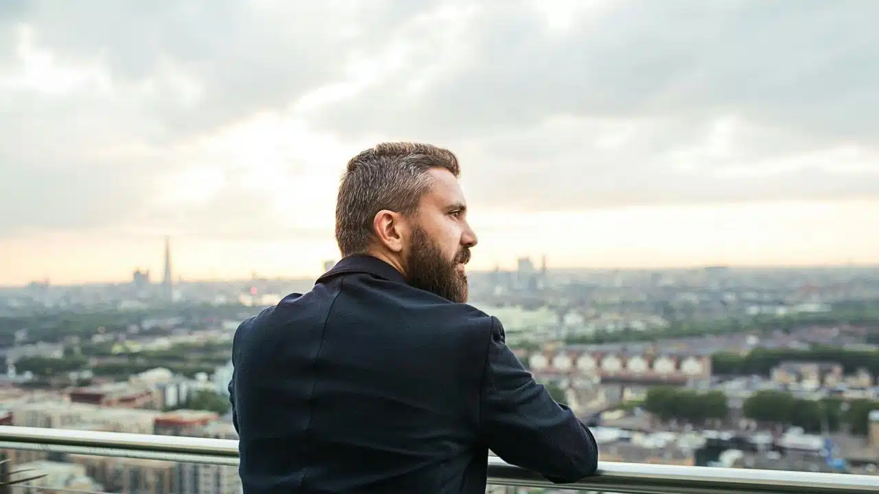 A bearded man leaning on a railing overlooking a cityscape.