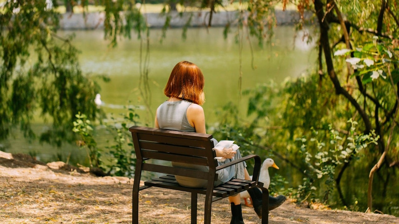 A woman reading a book while sitting on a bench next to a lake.
