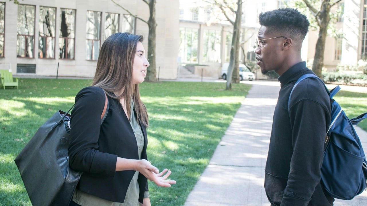 Two students, a woman and a man, talking outdoors on a university campus path.
