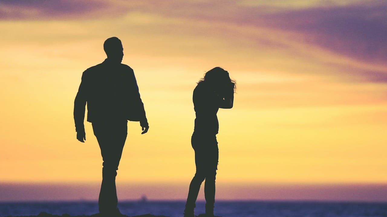 A silhouette of a couple walking into the sunset while the woman looks flustered.