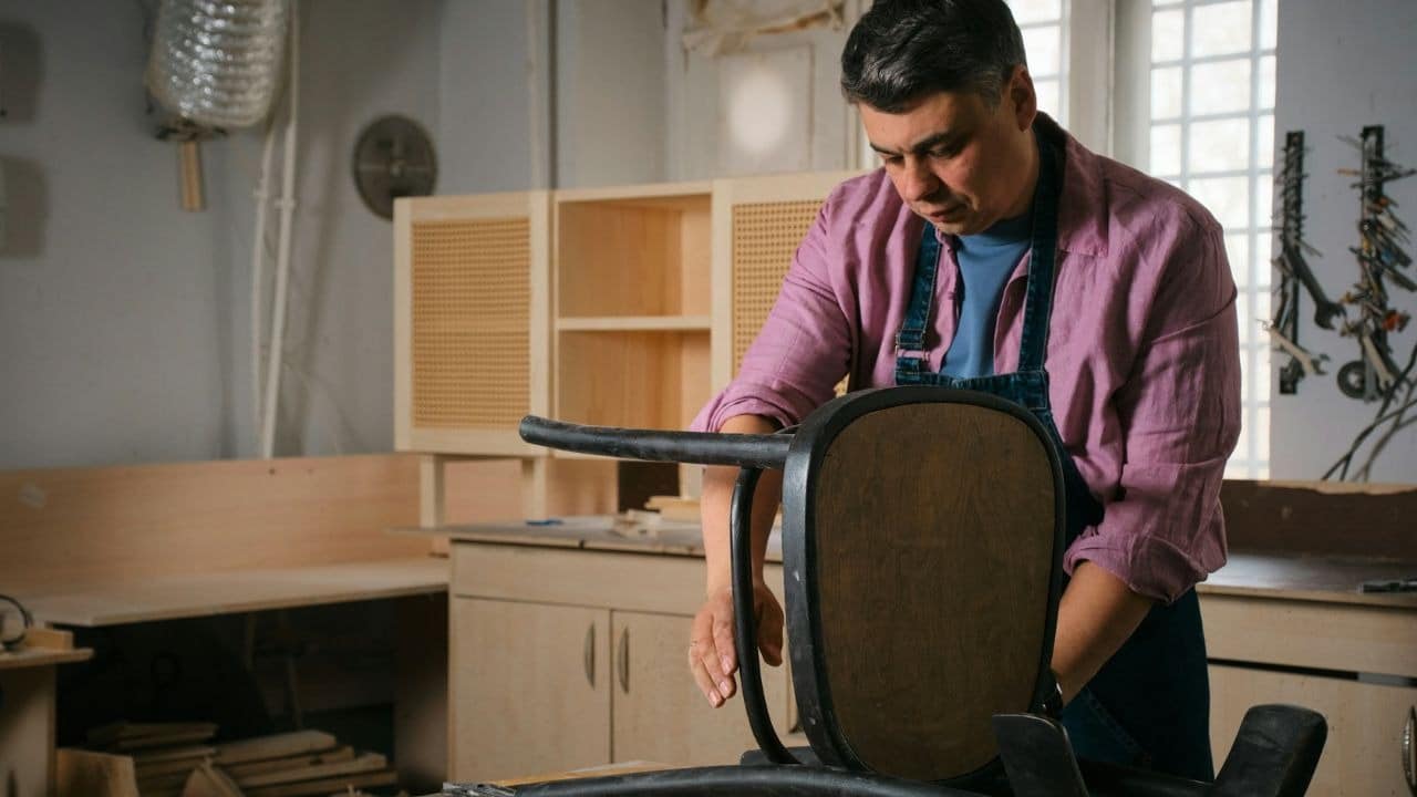 A craftsman in a pink shirt and apron works on repairing an upside-down wooden chair in a workshop.