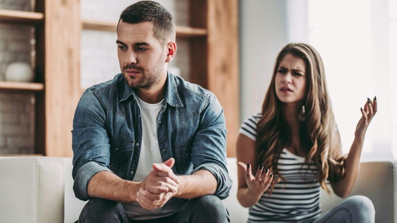 A man sits with a pensive expression while a woman behind him gestures with her hands.