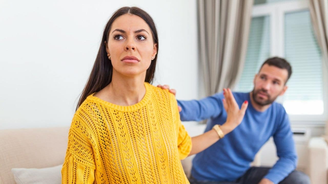 A man reaches out to a woman, who looks away from him with a pained expression and gestures for him to stop.