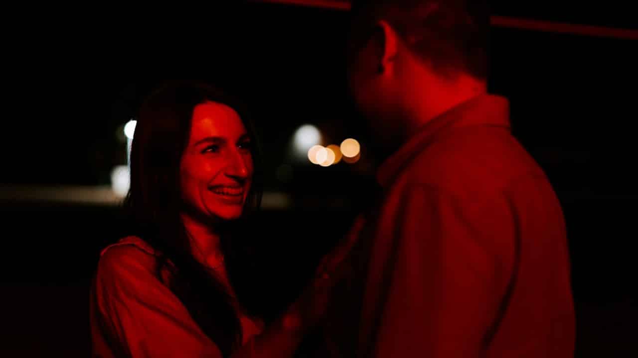 A smiling woman is looking up at a man in a dimly lit, red-hued setting.