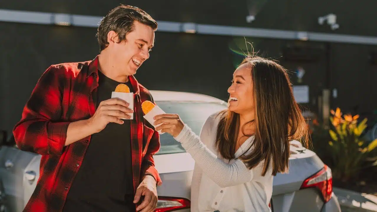 A couple is smiling at each other while standing by a car, holding and eating fried pies or hash browns.