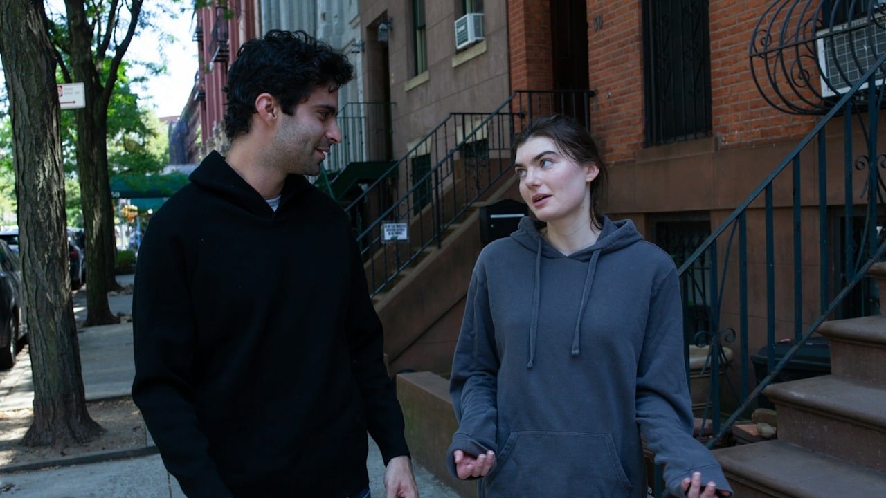 A man and a woman in hooded sweatshirts are walking and talking on a city sidewalk next to a brownstone building.