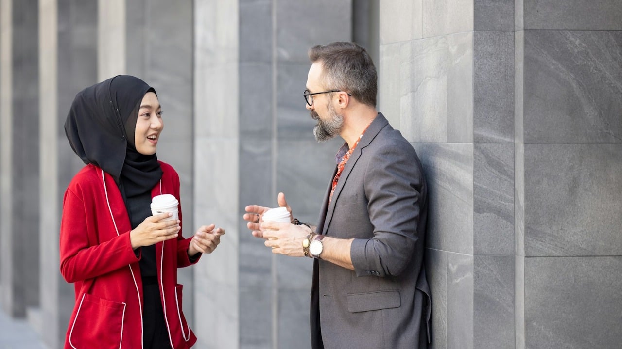 A woman wearing a hijab and a red blazer is standing and smiling while talking to a casually dressed man leaning against a stone column, both holding coffee cups.