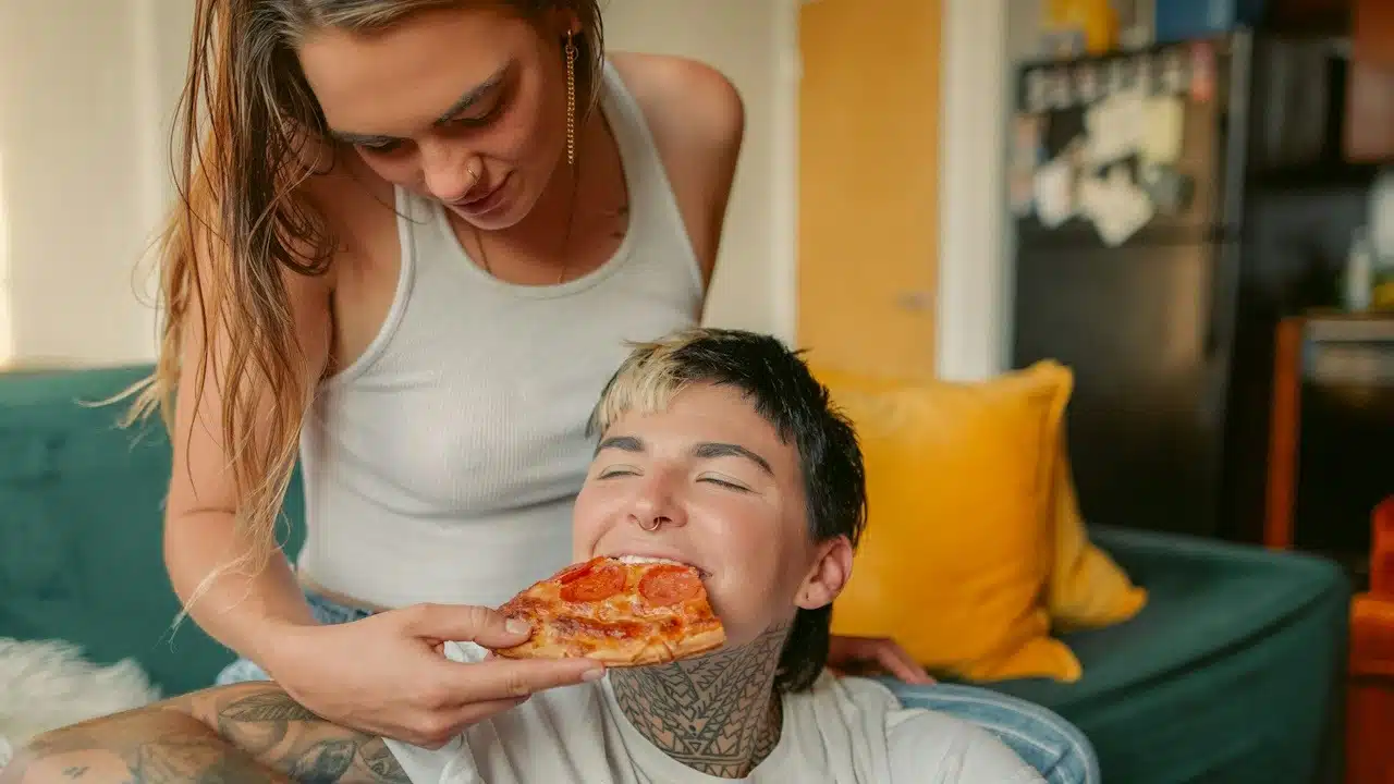 A woman sitting on a couch feeding a slice of pizza to a man who sits while leaning against her legs.