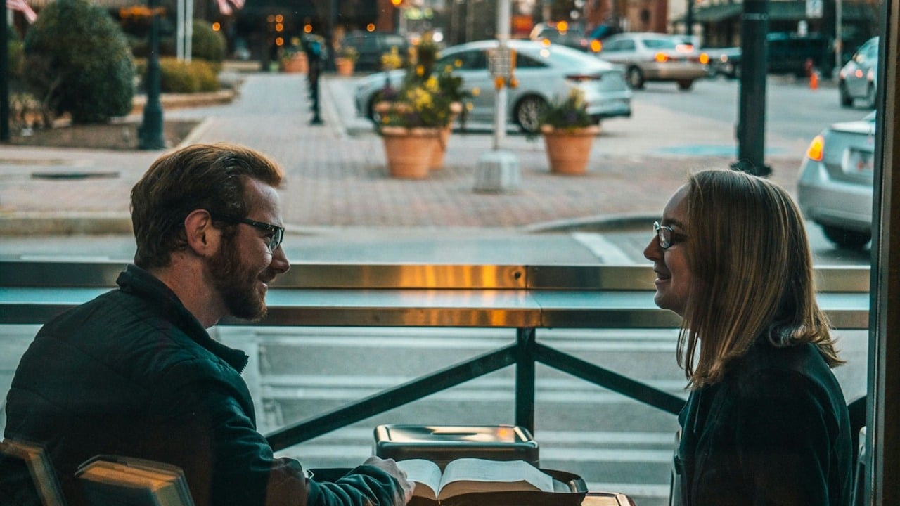 A man and a woman wearing glasses are sitting at a table by a window, looking at each other and smiling, with a book open between them.
