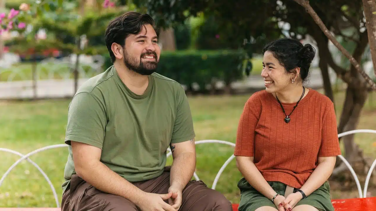 A smiling man and woman are sitting next to each other on a bench in a park, engaged in conversation.