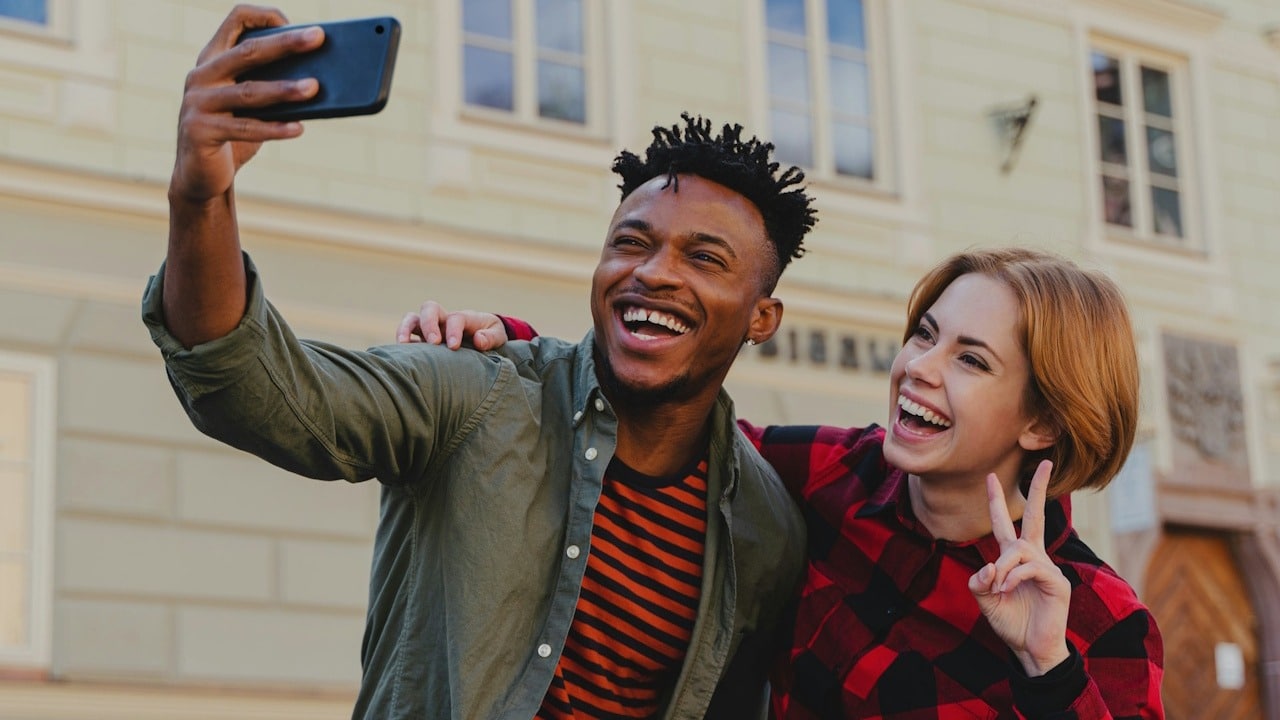 A joyful young man and woman are taking a selfie together outdoors in front of a building.