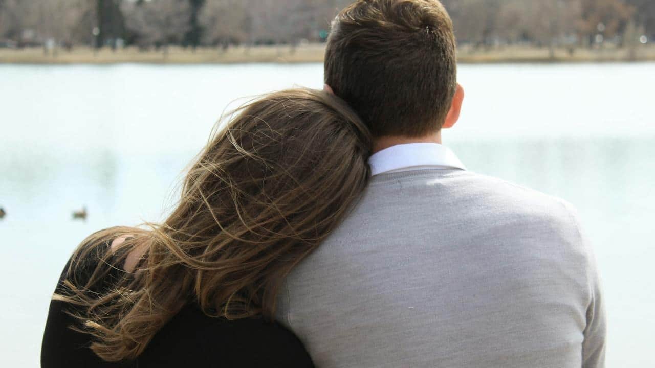 A couple sitting by the water with the woman resting her head on the man’s shoulder.