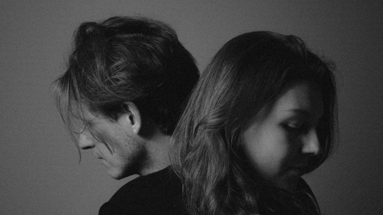 A couple standing with their backs to each other and gazing downwards.