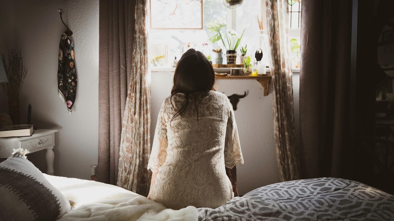 A woman sitting on a bed, facing away from the camera and looking out a brightly lit window.