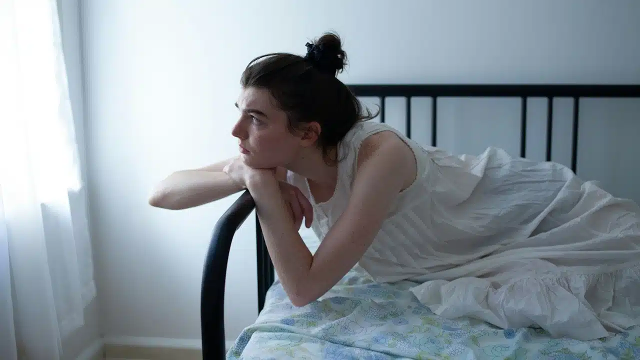 A young person in a white nightgown lying on a bed, looking thoughtfully out a bright window.
