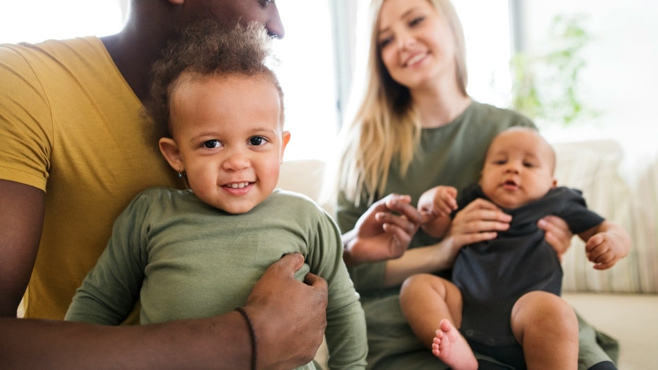 A man and woman sit on a couch together holding two small, smiling young children.