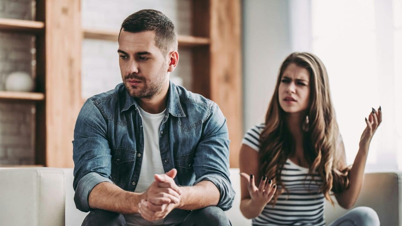 A serious man sits while a woman gestures angrily and speaks in the background.