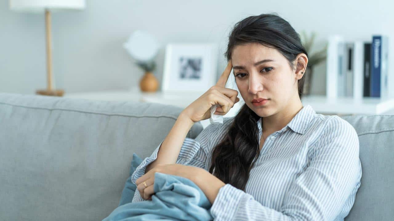 A woman sitting on a couch looking sad while holding a tissue.