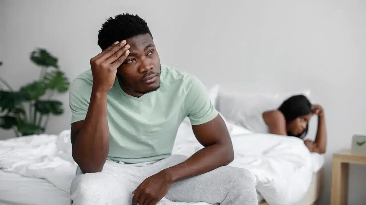 A man sits on a bed looking stressed with his hand on his forehead, while a woman lies facing away from him in the background