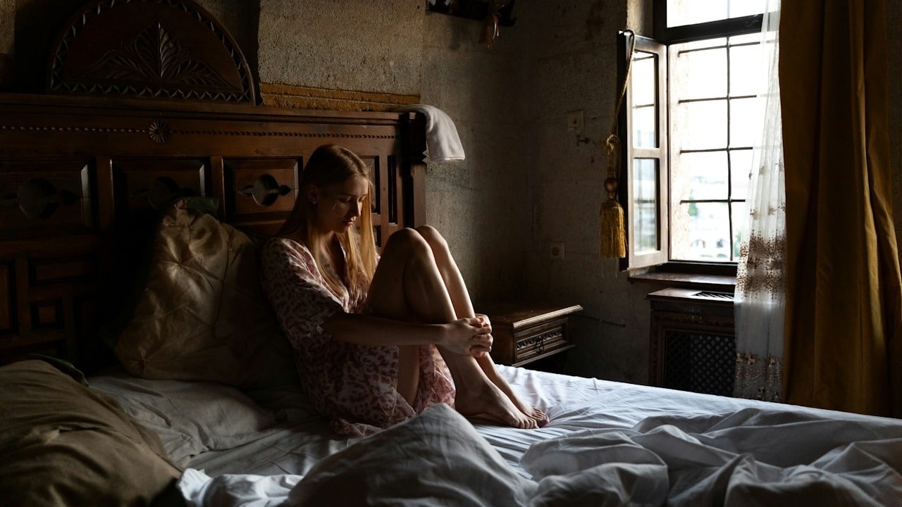 A lonely woman sitting hunched on a bed in a dark, old-fashioned room with a bright window.