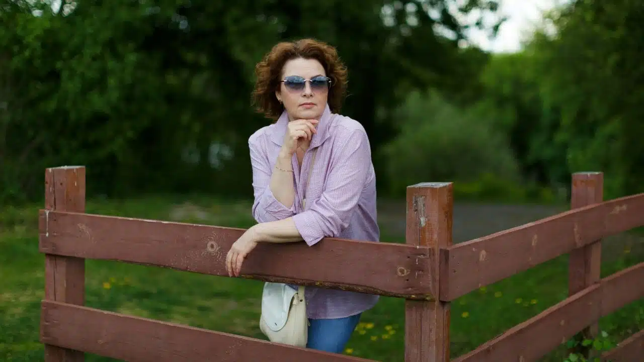 A confident woman with sunglasses is leaning on a wooden fence, looking thoughtful.