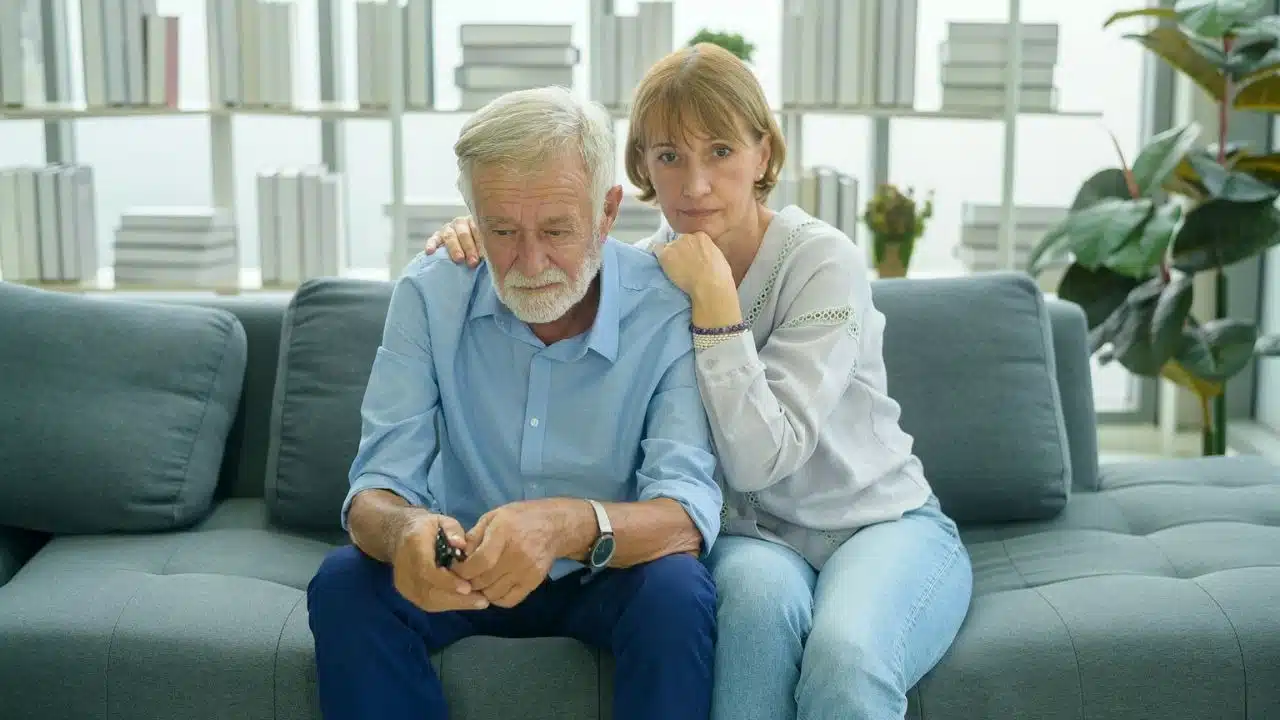 Elderly couple sitting sadly on a couch; woman's arm around man; bookshelf background.