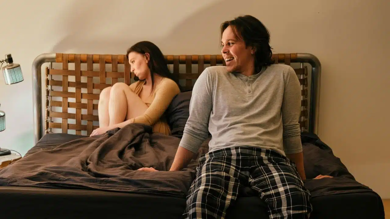 A woman sits with her back to a man who is smiling and laughing on a bed.