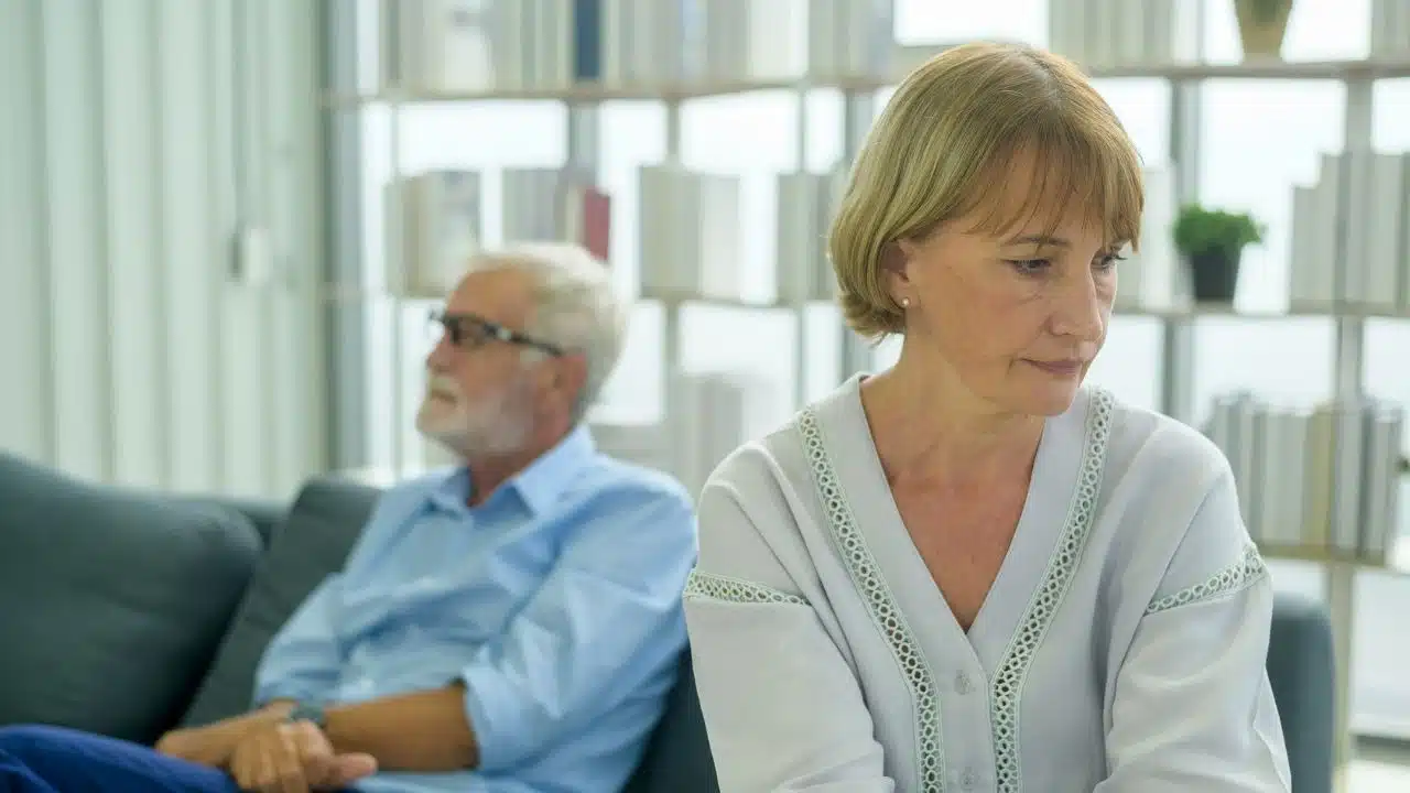 A sad, older woman sits on a couch in the foreground, with her husband visible but out of focus in the background.