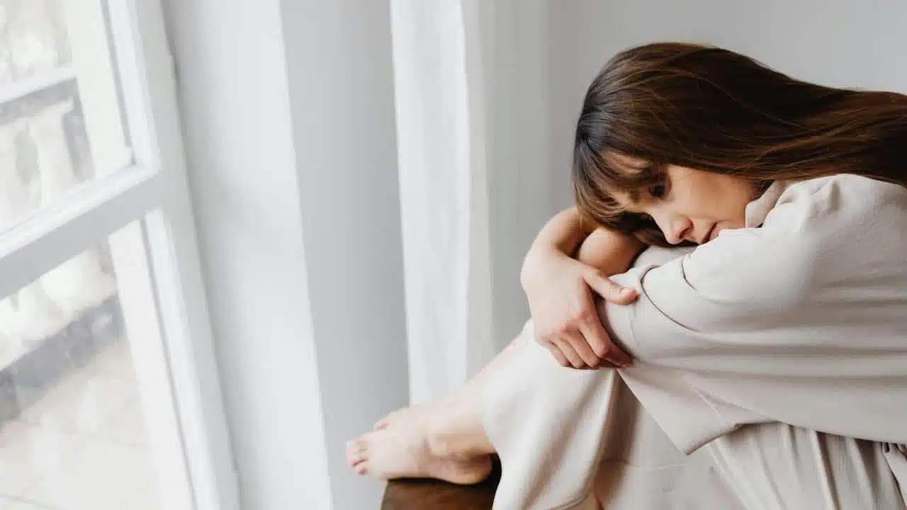 A woman sitting by a window, hugging her knees and appearing sad.