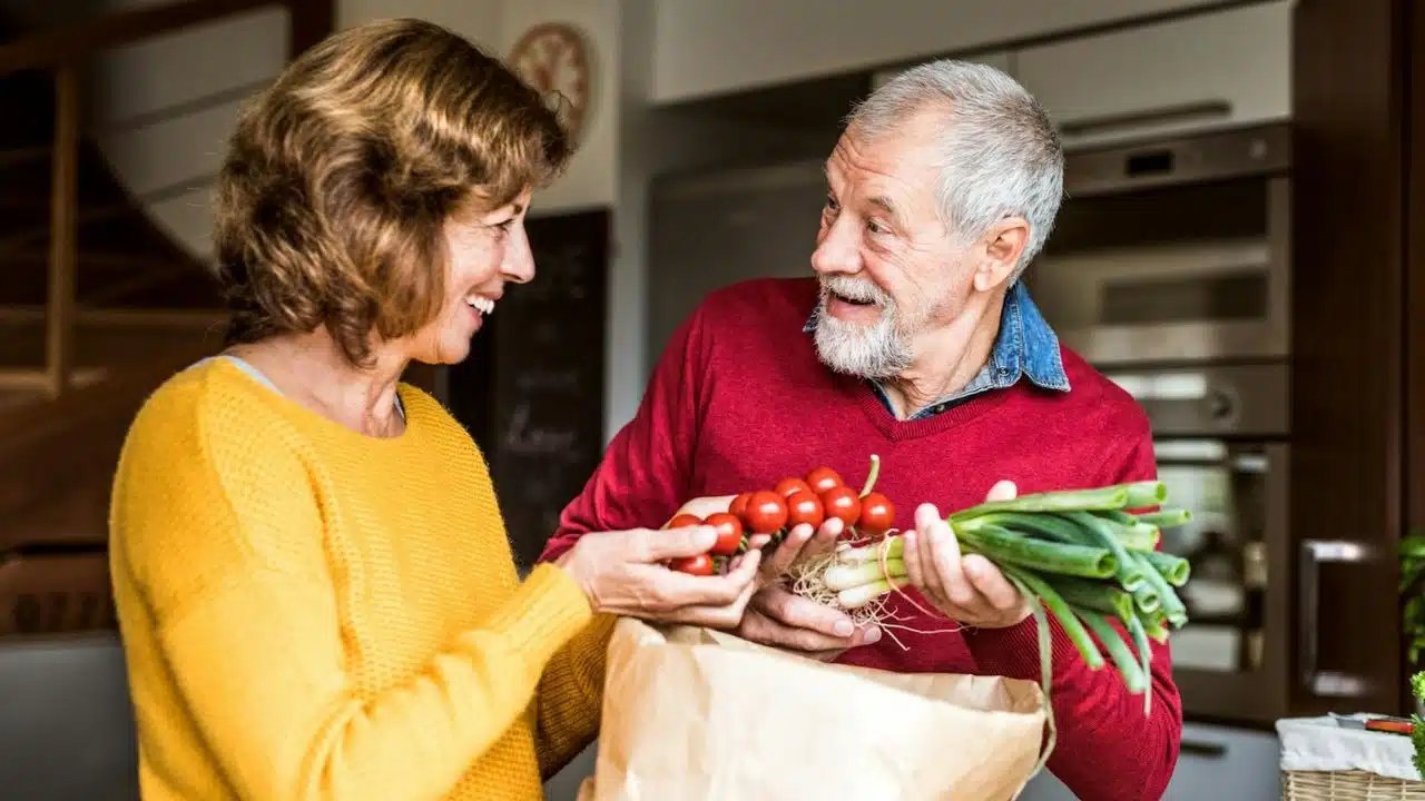 Happy older couple unpacking groceries, holding scallions and cherry tomatoes in a kitchen.