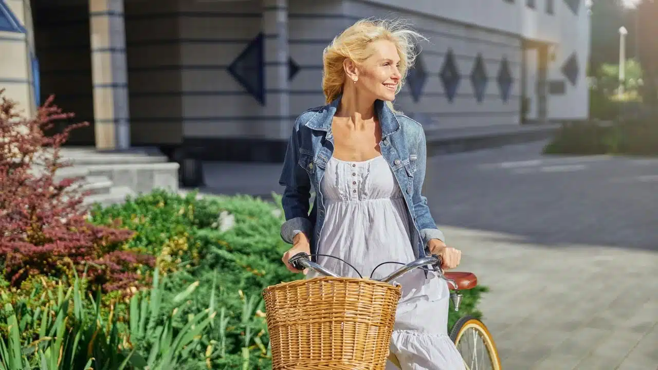 A smiling, mature woman in a denim jacket and white dress with a bicycle.