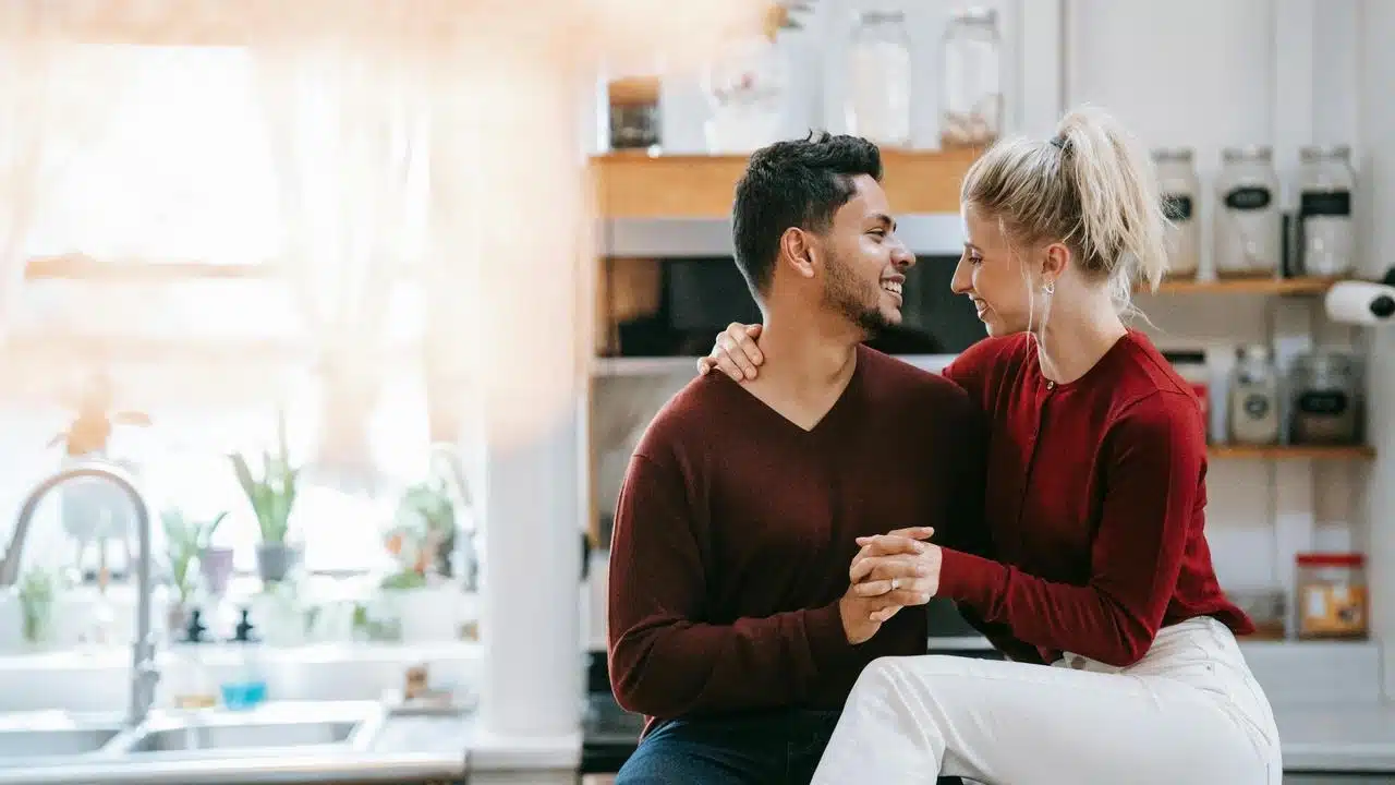 A couple sitting together in a kitchen, smiling and holding hands.
