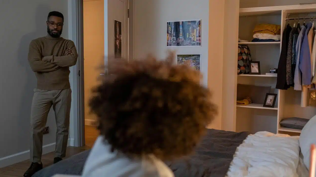 A man standing with crossed arms in a bedroom looking at someone sitting on the bed.
