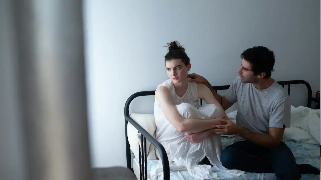 A man sitting next to his angry wife on bed.