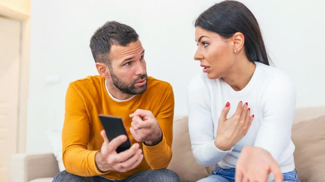 A man in an orange shirt holds a phone while talking to a gesturing woman.