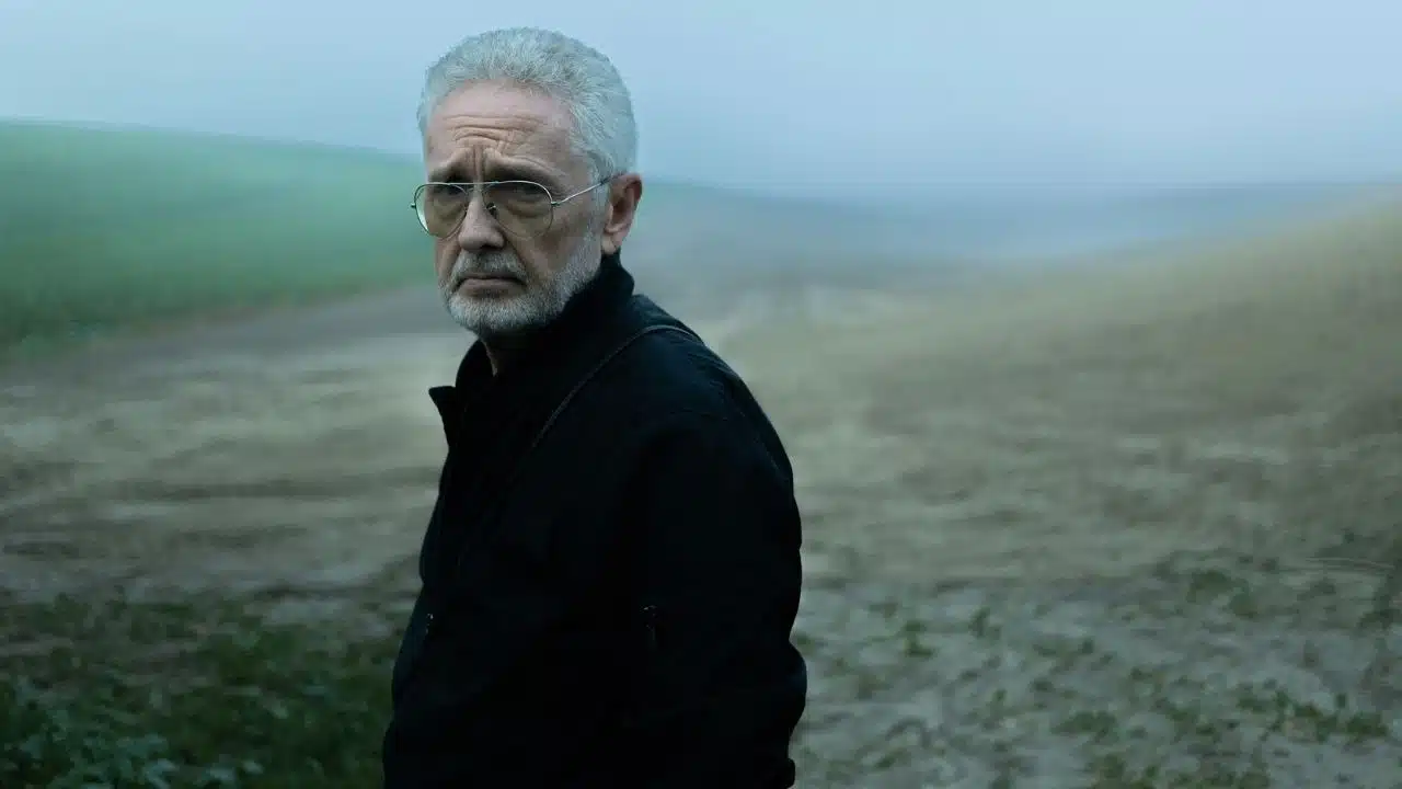 A senior man with glasses and a beard stands in a misty, rural field.