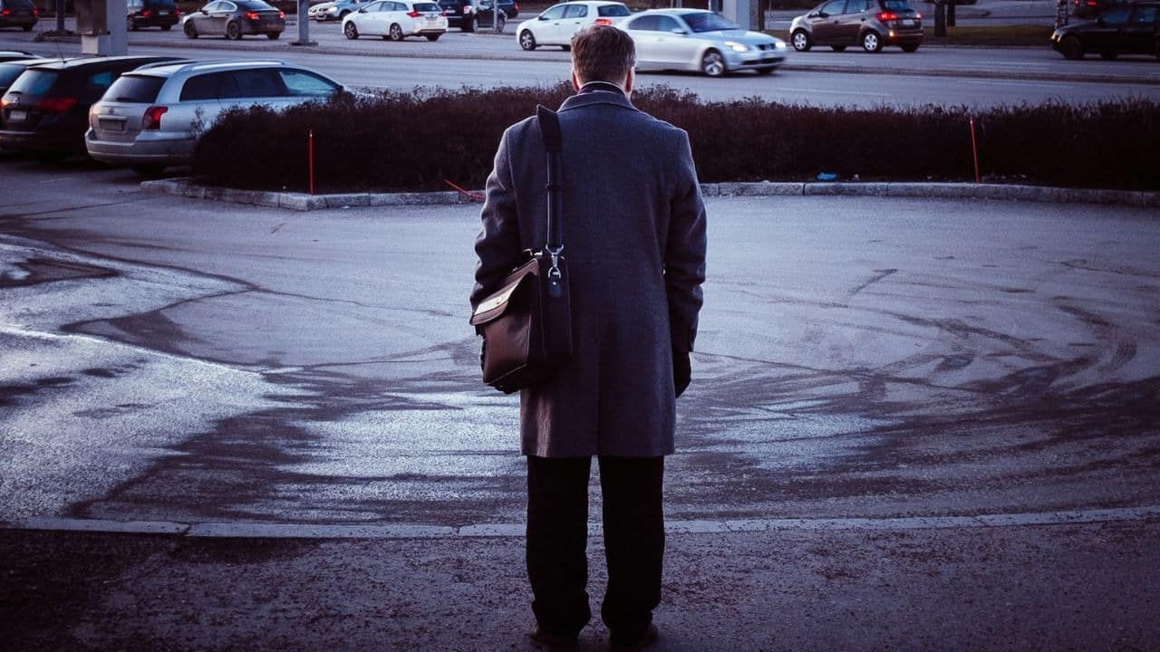 A man in a coat and briefcases stands in a parking lot, watching cars pass.